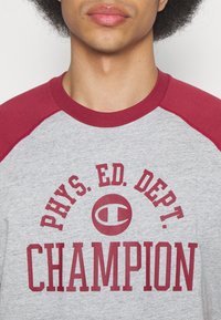 Camiseta gris de punto con mangas raglán rojas. Presenta un texto en negrita: “DEPARTAMENTO DE EDUCACIÓN FÍSICA CAMPEÓN” con un logo circular. Textura suave.