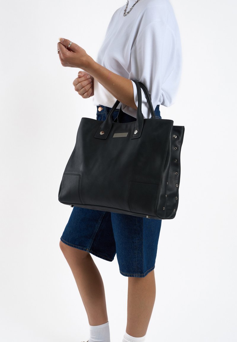 Zwarte leren tote bag met dubbele handvatten, vlakke voorzakken en decoratieve hardware accenten aan de zijkanten. Glad, gestructureerd ontwerp.