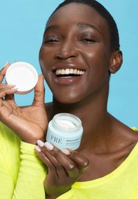 Un pot en verre bleu clair étiqueté "FRÉ MOISTURIZE ME", rempli de crème blanche, tenu par une personne portant un haut jaune vif et souriant.