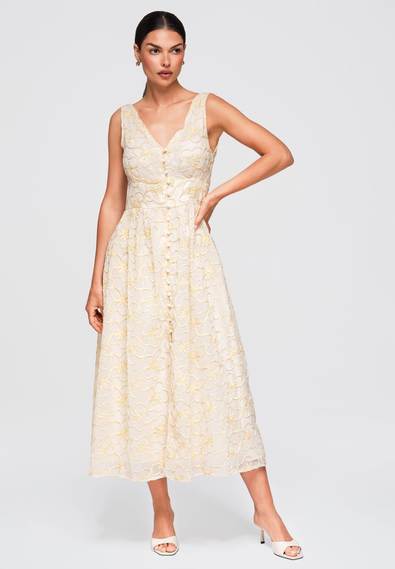 Femme portant une robe midi crème sans manches avec broderie florale jaune et boutons sur le devant, associée à des talons blancs à bout ouvert.