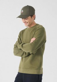 Joven con gorra verde oliva y sudadera a juego, con los brazos cruzados, sonriendo y mirando hacia abajo frente a un fondo claro.