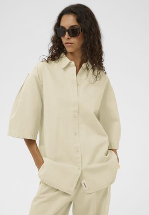 Femme aux cheveux bouclés portant une chemise boutonnée beige oversize et un pantalon assorti, avec des lunettes de soleil rectangulaires noires, mains dans les poches.