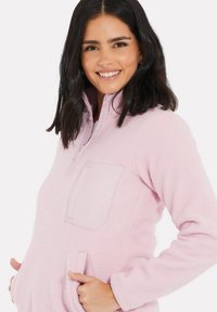 Pull-over en polaire rose clair avec un col zippé, doté d'une poche avant et de deux poches latérales. Texture douce et coupe décontractée.