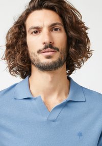 Helles blaues Polohemd aus weichstem Material. Es hat einen Kragen, kurze Ärmel und ein dezentes Logo, das auf der Brust gestickt ist.