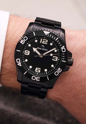 Montre de plongée noire avec une finition mate, affichant un cadran rond noir, des marqueurs d'heure lumineux et un bracelet en métal. Logo à 12 heures.