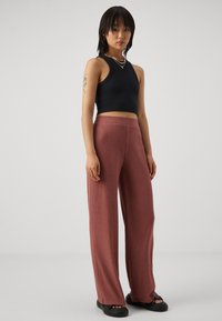 Schwarzes ärmelloses Top kombiniert mit weit geschnittenen, gerippten bordeauxroten Hosen. Das Modell trägt schwarze Slipper und eine mehrfarbige Perlenkette.