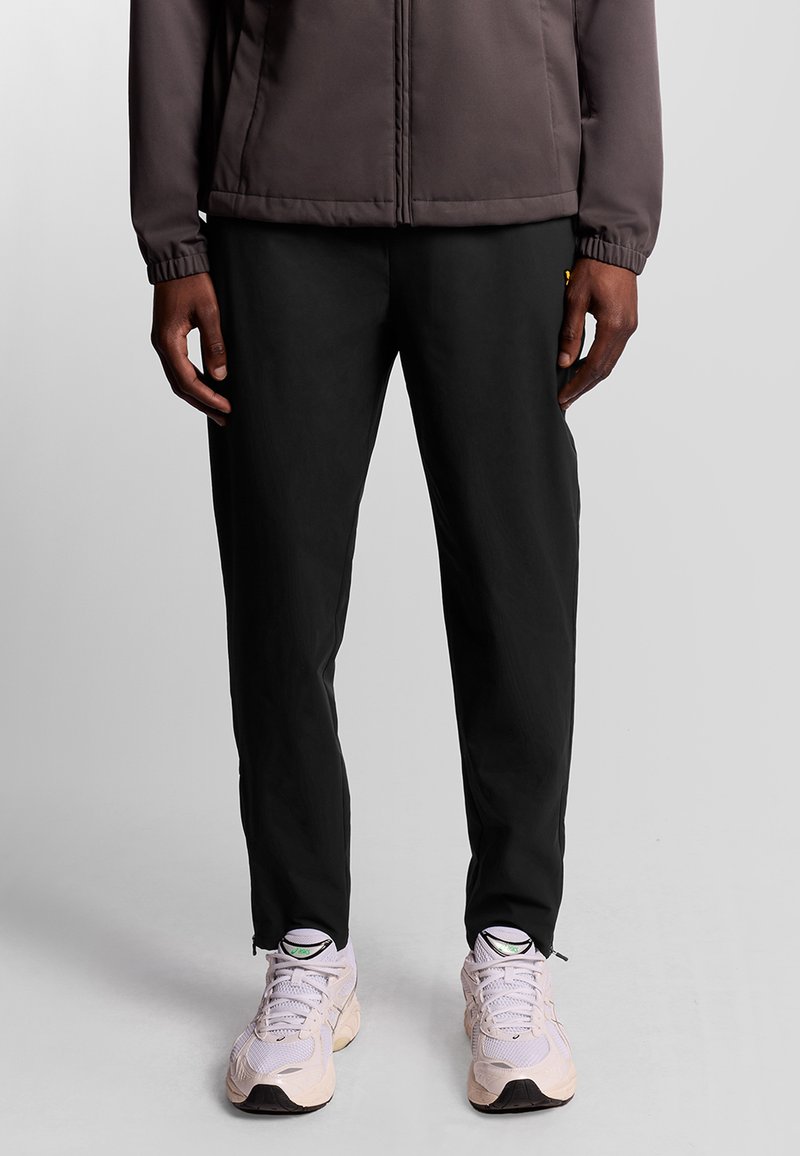 Zwarte broek van lichtgewicht materiaal, met een rechte snit en zijrits onderaan, gecombineerd met lichtgekleurde sneakers.