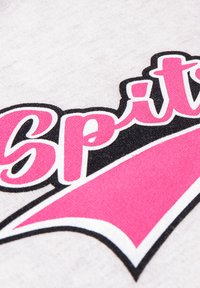 Witte stof met een textuurpatroon waarop het woord "Spit" in roze en zwart is afgebeeld, aangevuld met een roze grafische pijl.