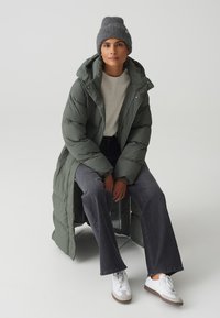 Lange groene pufferjas met een hoge kraag, grijze gebreide beanie, crème trui en donkergrijze wijde broek. Witte sneakers met bruine accenten.