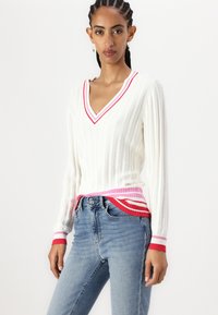 Maglione bianco a coste con scollo a V profondo, caratterizzato da accentuazioni a strisce rosa e rosse sui polsini e sull'orlo, abbinato a jeans blu.
