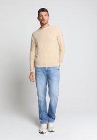 Beige trui met lange mouwen en een gestructureerd ontwerp, gecombineerd met lichtblauwe denim jeans. Het model draagt witte en grijze sneakers.