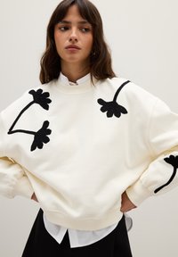 Felpa bianca con ricami floreali neri e vestibilità rilassata. Presenta polsini e orlo a costine, sovrapposta a una camicia bianca con colletto.