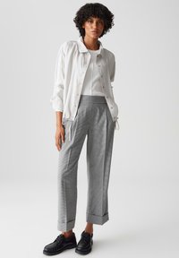 Witte lichte jas, aansluitende grijs geruite broek en zwarte schoenen. De jas heeft een knoopsluiting; de broek heeft een omslag.