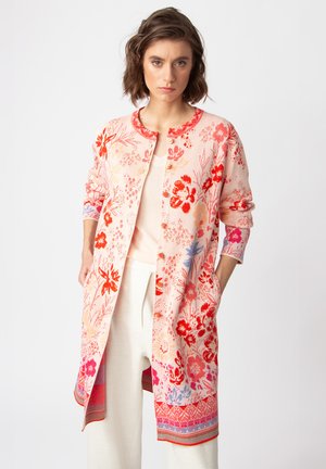 Lange roze cardigan met rode bloemenpatronen, ronde halslijn en knoopsluitingen. Heeft zakken en kleurrijke afwerking aan de onderrand en manchet.