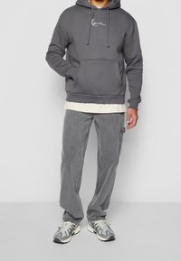 Felpa grigia con tasca frontale e logo bianco, indossata sopra una T-shirt color crema. Abbinata a pantaloni cargo grigi e sneakers multicolori.