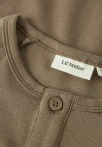 Ruda medvilninė marškinėlių apykaklė su mygtuko uždarymu, siuvinėta etikete "Li'l'Atelier" ir lygiu, raukšlėtu tekstūru.