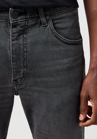 Nærbilde av svarte denimjeans med frontlomme, knapp, glidelås og en mørkhudet hånd avslappet på siden.