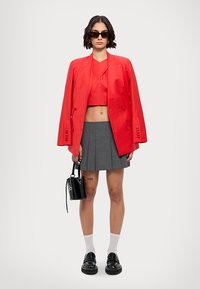 Červený saténový blazer s krátkým střihem přes červený top, šedá skládací sukně, černá taška a robustní černé boty. Modelka nosí sluneční brýle a bílé ponožky.