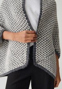 Bouton de fermeture à la main sur un cardigan en tricot texturé gris et blanc porté sur un haut blanc et un pantalon noir.
