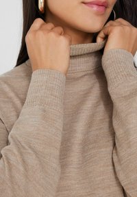 Suéter beige de cuello alto con puños acanalados, textura suave y un cuello ajustado. Manos tirando del cuello hacia arriba.