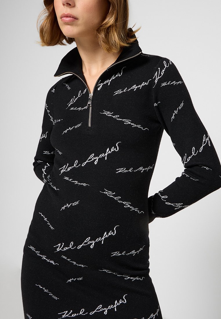 Femme portant une robe noire à manches longues avec un texte en cursive blanche et une fermeture éclair à l'avant du col, se tenant les mains derrière le dos.