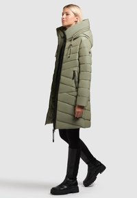 Olijfgroene pufferjas met een hoge kraag, zipzakken en een verstelbare capuchon. Heeft een gewatteerd patroon en zwarte accenten.