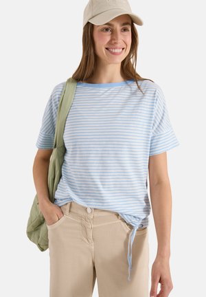 Femme souriante, portant une casquette beige, un haut à manches courtes rayé bleu clair et blanc avec lien de serrage, un pantalon beige, et portant un sac matelassé vert sur l'épaule.
