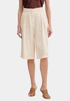 Donna che indossa pantaloncini plissettati color crema fino al ginocchio, top marrone senza maniche e mule intrecciate marrone chiaro con tacco, in piedi su sfondo bianco.