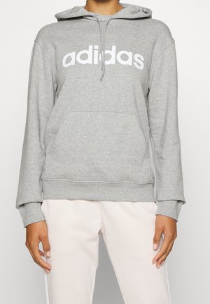 Person trägt einen grauen Adidas Hoodie mit weißem Logo und weißen Hosen, steht mit entspannten Armen an den Seiten.