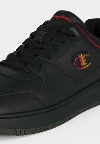 Baskets basses noires avec lacets noirs, coutures rouges et logo Champion rouge et jaune sur le côté.