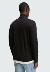 Donker gebreide trui met een hoge kraag, geribde manchetten en zoom, met een zachte textuur en een aansluitend ontwerp. Gedragen over een wit shirt.