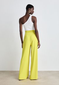 Wijde broeken in felgeel, gemaakt van een gladde stof met een hoge taille. G gedragen met een strakke witte top met één schouder.