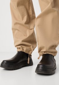 Black rubber boots with a chunky sole and rounded toe, paired with beige elastic cuffed pants. Matte finish on boots, textured fabric on pants.