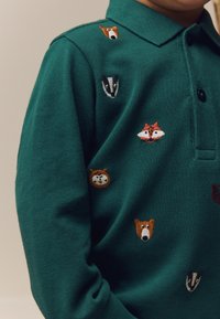 Žalia polo marškinėliai iš faktūrinės medžiagos su siuvinėtais gyvūnų piešiniais: lokys, lapė, barsukas ir animacinio stiliaus figūros įvairių spalvų.