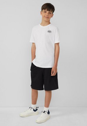 Wit katoenen t-shirt met een klein zwart logo op de borst, gecombineerd met zwarte shorts en witte sneakers met marineblauwe accenten.