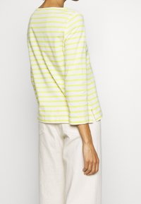Frau, die ein weißes Langarmshirt mit hellgrünen horizontalen Streifen und off-white Hosen trägt, von hinten vor einem schlichten Hintergrund gezeigt.