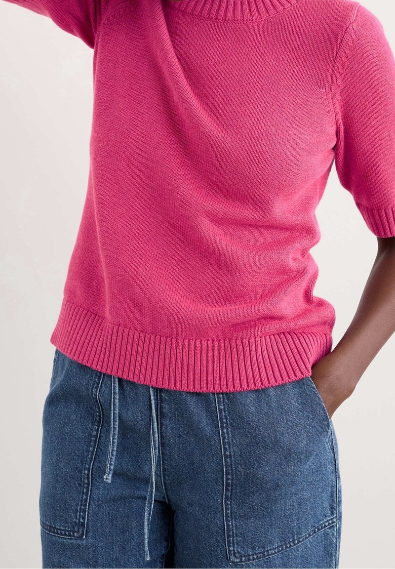 Fuchsia short-sleeve pullover in soft knit with a ribbed neckline and hem, paired with loose-fitting denim trousers featuring an elastic waistband.