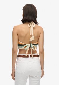 Gehaakte haltertop in oranje, groen en crème. Heeft een strik aan de achterkant met franjes, gedragen met witte high-waisted jeans en een gevlochten bruine riem.