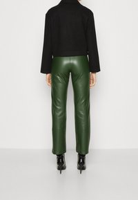 Veste noire cropped avec des manches longues, associée à un pantalon en cuir vert moulant et des chaussures à talons noirs. Textures lisses, design ajusté.