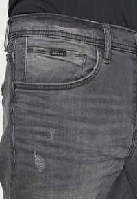 Jeans de dril gris con un aspecto desgastado, que cuentan con múltiples bolsillos, una superficie texturizada y marcas de desgaste sutiles cerca del bolsillo trasero.
