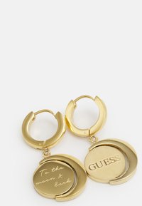 Boucles d'oreilles en or avec des charms gravés : l'un porte l'inscription "À la lune et retour" et l'autre a "GUESS" avec une finition lisse et brillante.