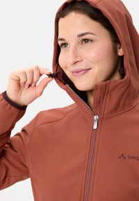 Woman adjusting the drawstring on the hood of a rust-colored softshell jacket with a zipper and logo on the chest.