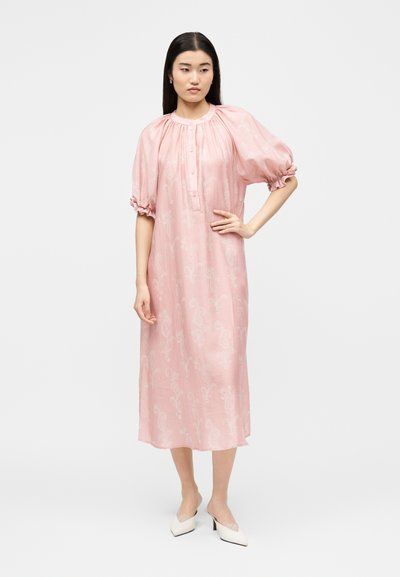 Γυναίκα που στέκεται φορώντας ένα χαλαρό, ανοιχτό ροζ μίντι φόρεμα με φουσκωτά μανίκια και άσπρα μυτερά παπούτσια μπροστά σε μια απλή φωτεινή επιφάνεια.