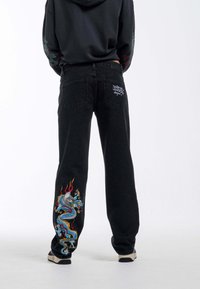 Jeans de mezclilla negra con un diseño de dragón bordado multicolor en la pierna izquierda, con un corte casual y bolsillos traseros estándar.