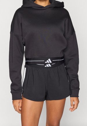 Osoba nosząca czarną skróconą bluzę z kapturem oraz czarne krótkie spodenki sportowe z czarnym pasem i białym logo Adidas.