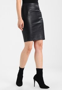 Jupe crayon en cuir noir au design ajusté, texture lisse et coutures renforcées. Association avec des bottines à talons hauts noires.