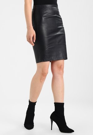 Jupe crayon en cuir noir au design ajusté, texture lisse et coutures renforcées. Association avec des bottines à talons hauts noires.