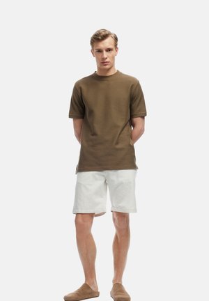 T-shirt marron à manches courtes côtelées associé à un short en lin blanc et des chaussures slip-on marron sur un fond blanc uni.