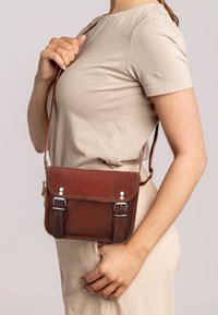 Brun læder crossover taske med sølvhardware, justerbar rem og klaplukning. Glat tekstur med spænde detaljer og kontrasterende syninger.