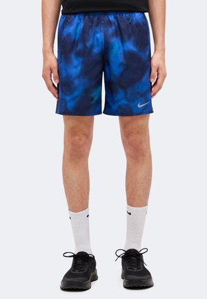 Mann trägt blaue und schwarze gemusterte Sportshorts mit weißen Sneakersocken und schwarzen Turnschuhen, steht mit entspannten Armen an den Seiten.